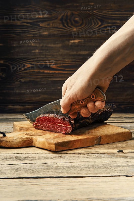 Hand with knife cutting smoked dried beef meat on slices on wooden cutting board