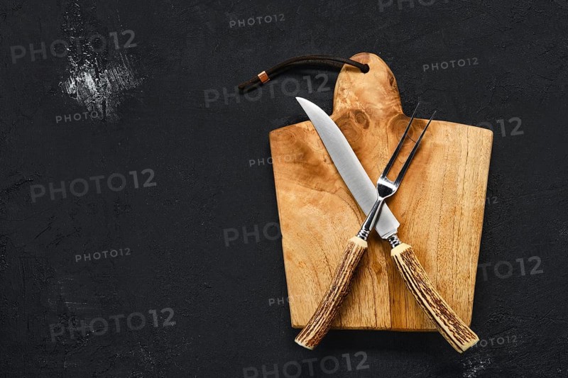 Overhead view of wooden cutting board with steak knife and fork on black shabby background