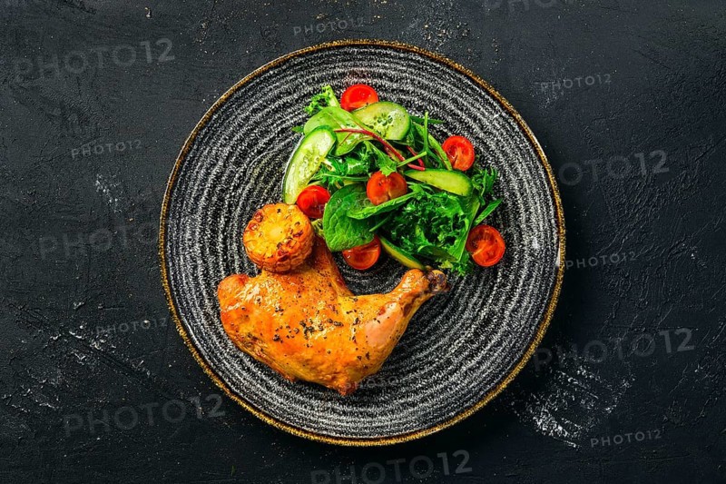 Top view of baked chicken thigh served with fresh vegetables and grilled corn on dark background