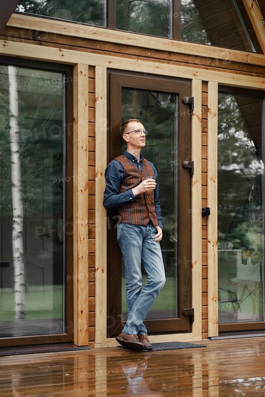 Pensive middle age man stands on the terrace of a wooden bungalow and drinks bourbon