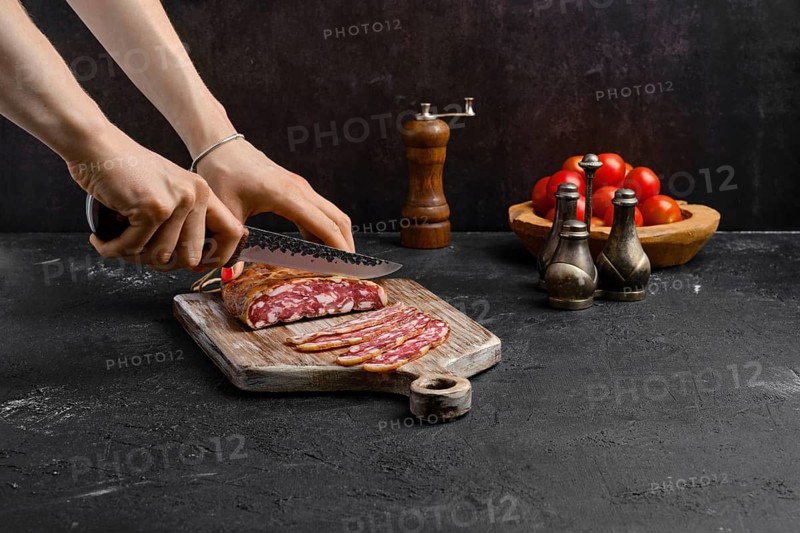 Female hand with knife slicing smoked pork sausage in organic casing on wooden cutting board