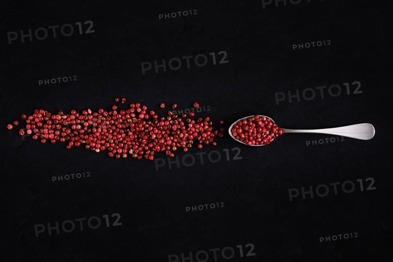 Dry pink pepper scattered near metal spoon on dark background