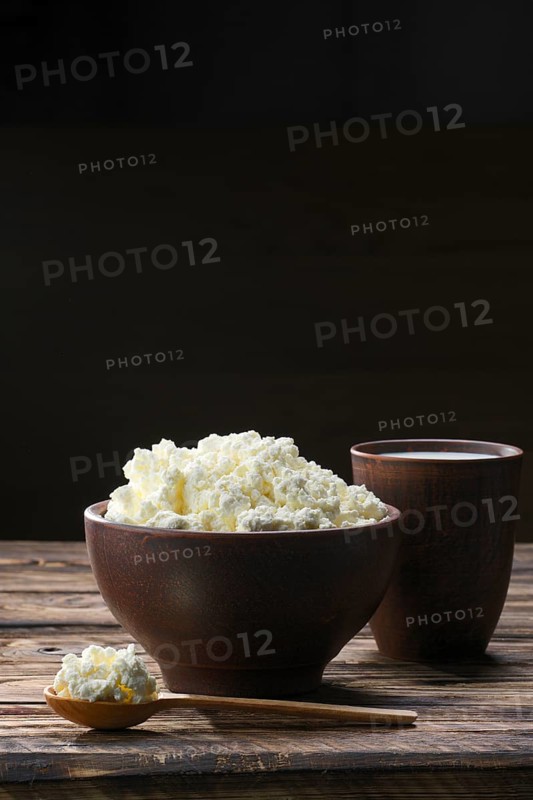 Fresh cottage cheese in clay bowl with wooden spoon with a glass of milk on rustic wooden background