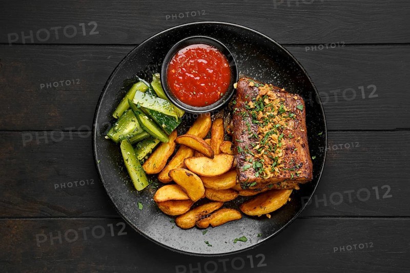 Top view of roasted beef ribs with potato wedges and pickled cucumber on a plate
