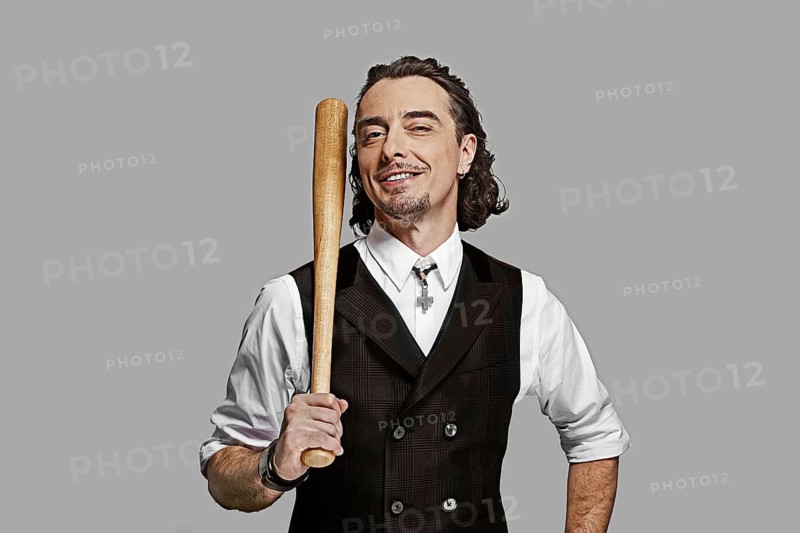 Dramatic man in white shirt and black vest with long curly hair holding baseball bat