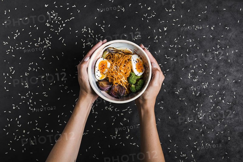 Elevated view hands holding bowls ramen noodles with eggs salad spread with rice grains black background