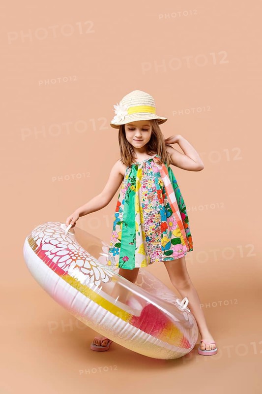 Cute child in sundress and straw hat with swimming ring playing in studio