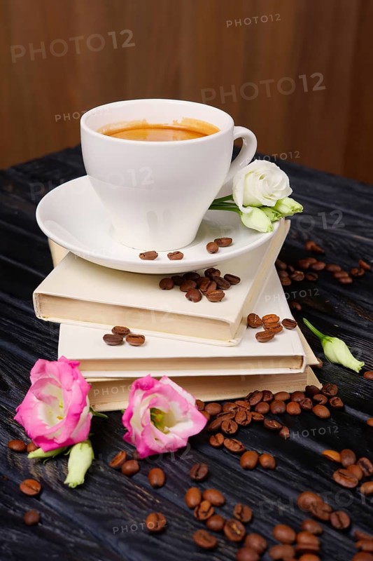 Cup of coffee on top of pile of books with coffee beans and flowers scattered on the table
