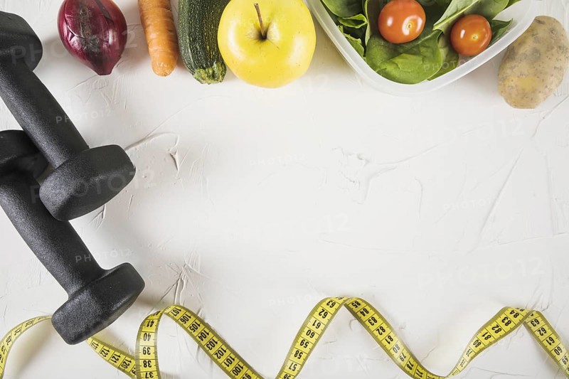 Overhead view of measuring tape food and dumbbell on white backdrop