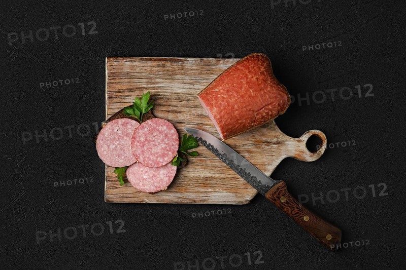 Overhead view of smoked beef salami and sandwich on cutting board over black background