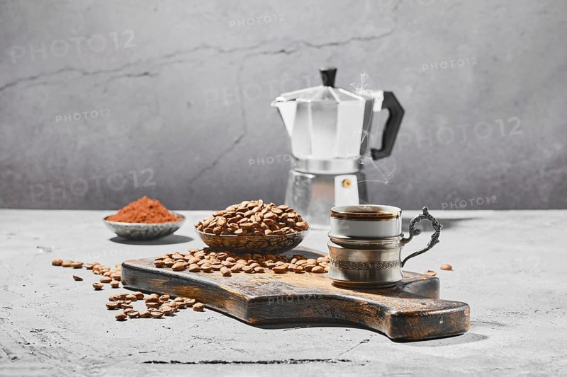 Hot cup of espresso with coffee beans scattered on wooden board and a pot under hard morning sun