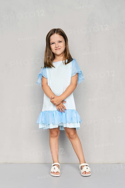 Cute little girl in blue dress and white sandals posing with hands clasped down near the gray wall