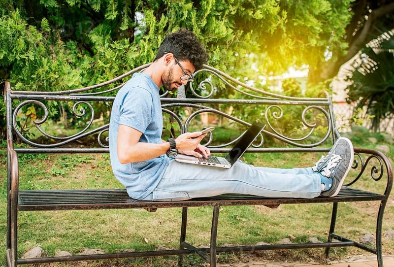 Freelancer man sitting in a park using laptop and cellphone. Man in a park working online with laptop. Relaxed man working with laptop outdoor
