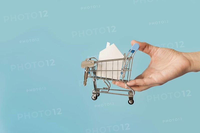 Close up hand holding miniature shopping cart with paper house keys against blue background