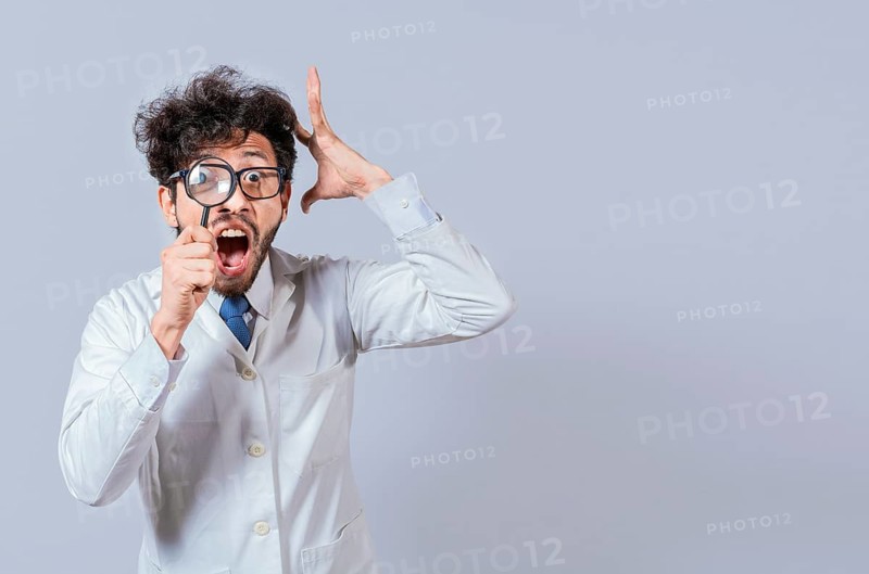 Surprised male scientist in lab coat looking at camera with magnifying glass. Mad scientist holding a magnifying glass isolated