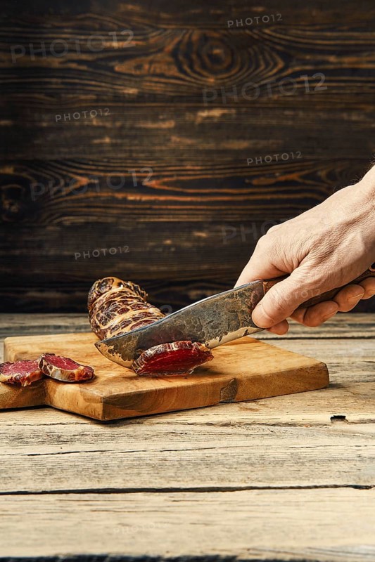 Hand with knife cutting smoked dried beef meat on slices on wooden cutting board