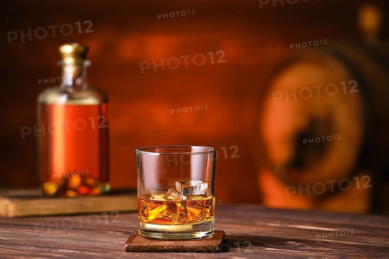 Glass of whiskey with ice on wooden table with bottle and barrel on background