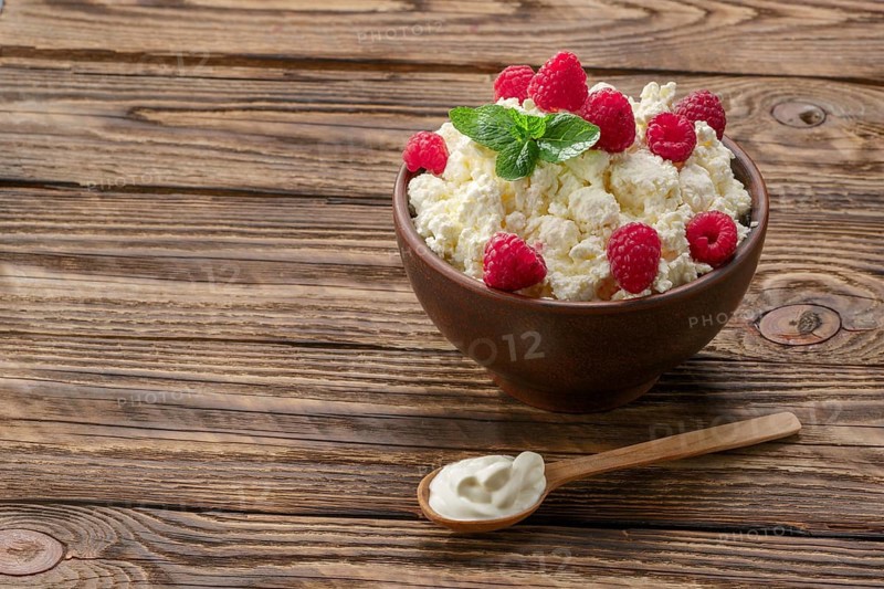 Wooden spoon with sour cream near clay plate with fresh cottage cheese with raspberries and mint