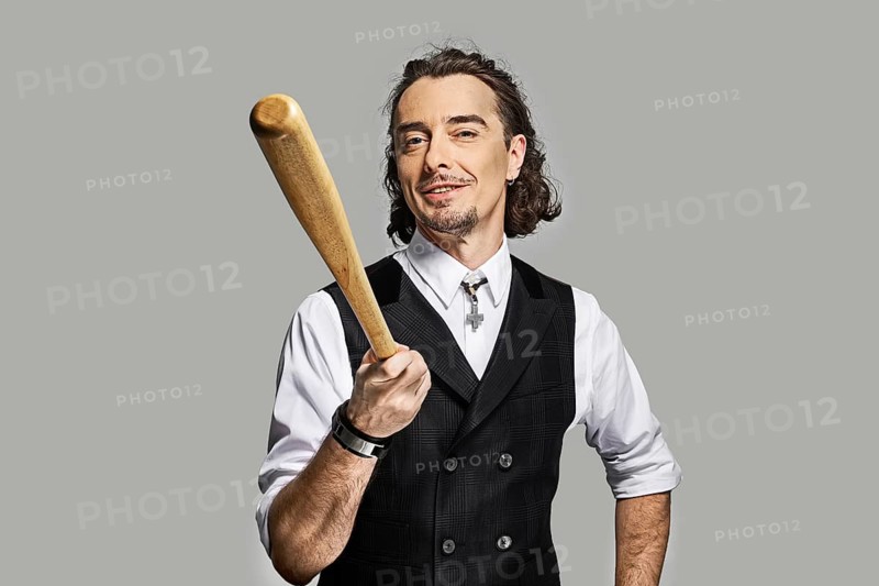 Dramatic man in white shirt and black vest with long curly hair holding baseball bat