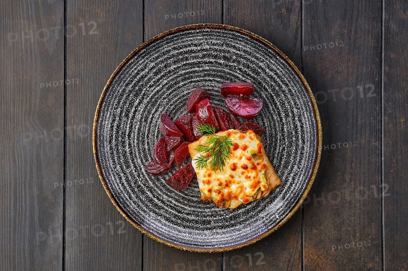 Top view of fried cod fillet with melted cheese topping and roasted beetroot slices on dark wooden table