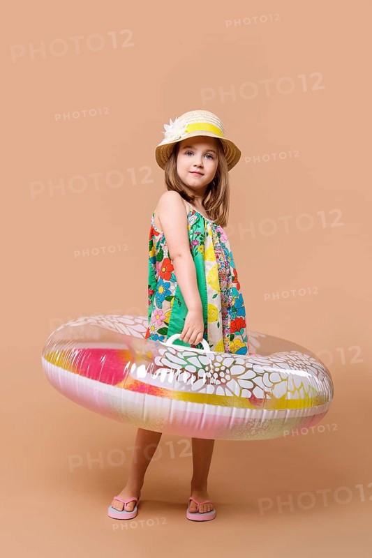 Cute child in sundress and straw hat with swimming ring playing in studio