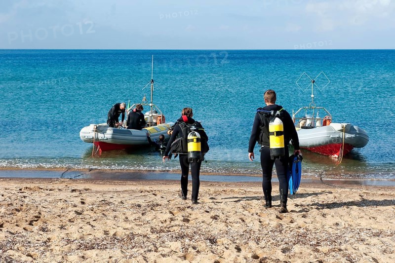 Divers Sport divers running with scuba equipment compressed air tanks across beach sandy beach from Mediterranean Sea to inflatable boat
