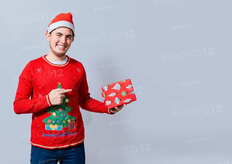 Smiling guy in christmas hat holding and pointing a gift box. Portrait of teenager in christmas hat holding and pointing a gift box. Guy in christmas hat and sweater holding a gift box isolated