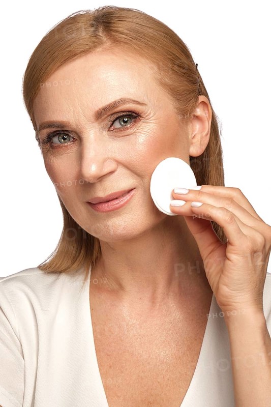Portrait of a beautiful elderly woman in a white shirt removing makeup with a cleanser and a cotton pad
