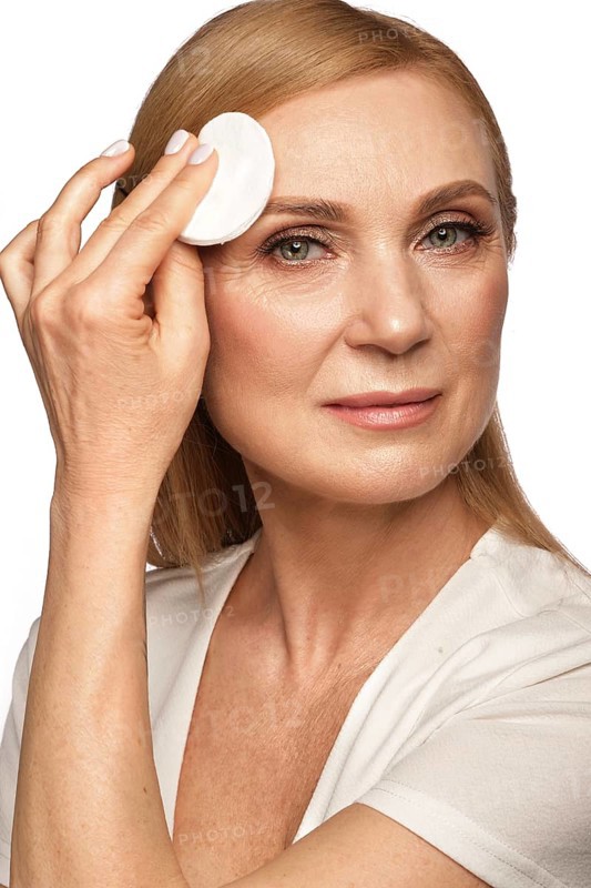 Portrait of a beautiful elderly woman in a white shirt removing makeup with a cleanser and a cotton pad