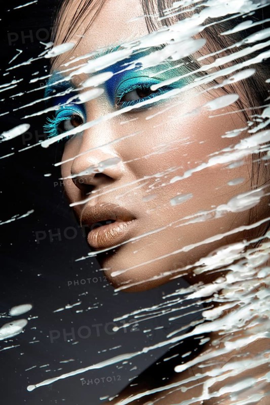 Beautiful Asian girl with bright blue make-up behind glass with drops of wax. Beauty face. Picture taken in the studio on a black background