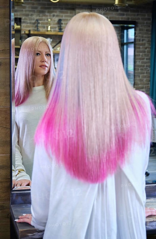 Beautiful blond girl with colored pink hair in a hairdressing salon. The client looks at herself in the mirror