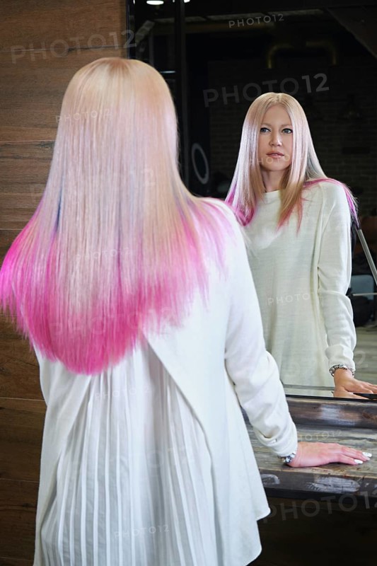 Beautiful blond girl with colored pink hair in a hairdressing salon. The client looks at herself in the mirror