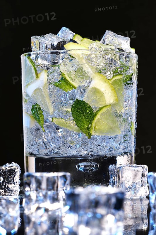 Glass of Mojito summer alcoholic cocktail with ice cubes mint and lime on black background with raw lime and mint leaf