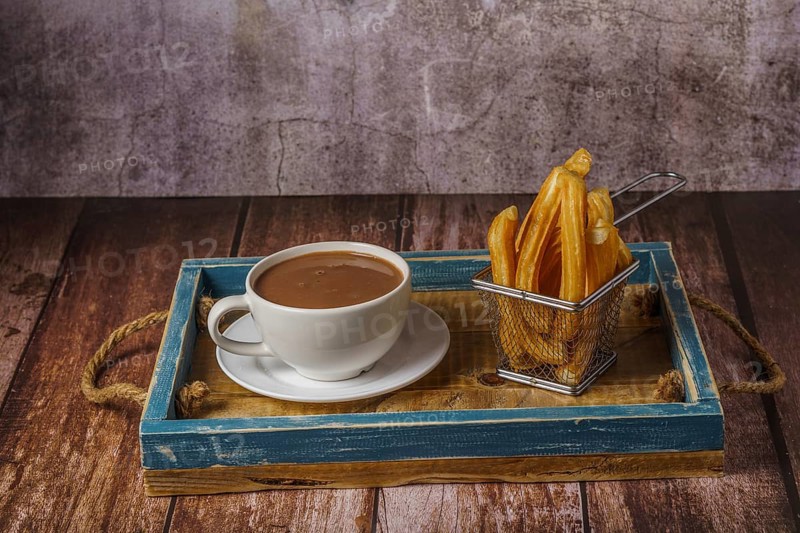 Hot chocolate in a white mug with churros in a blue wooden tray on a wooden table