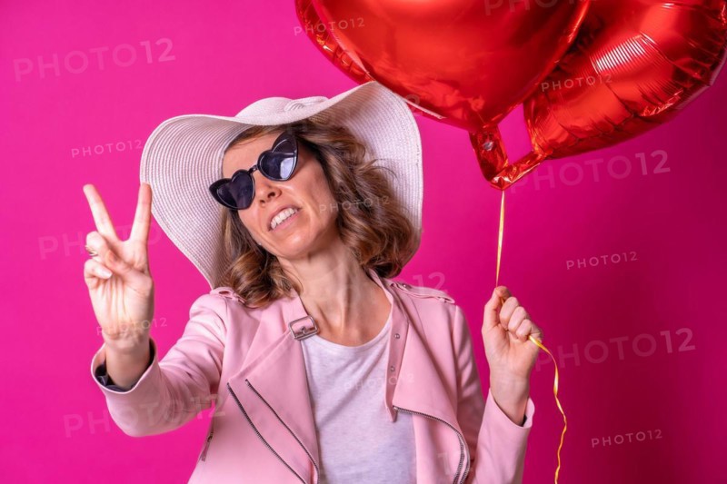 Blond caucasian woman in a white hat and sunglasses making the victory sign with her hands in a nightclub with some heart balloons