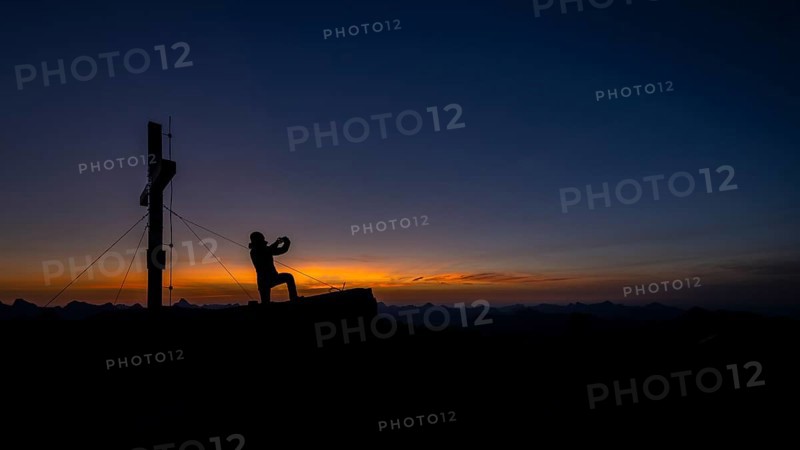 Mobile phone photographer at the summit cross of the Namloser Wetterspitze at sunset with Lechtaler Alpen