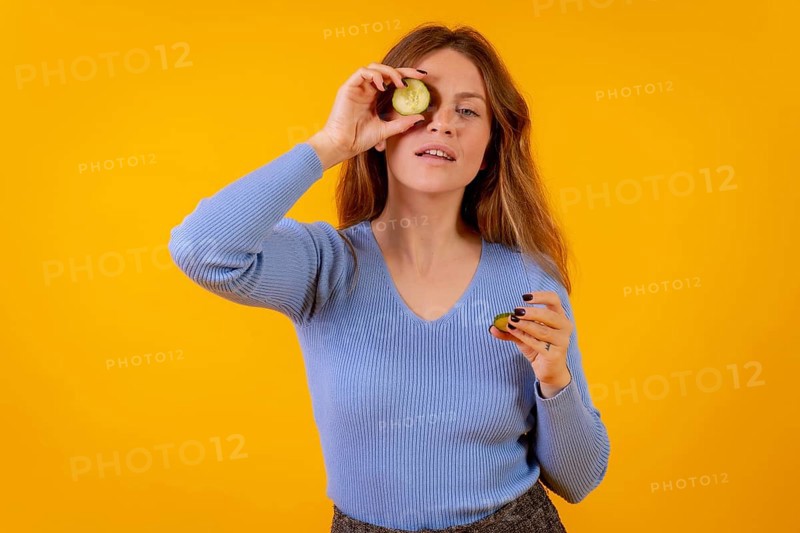 Vegan woman with a slice of cucumber on her eye on a yellow background