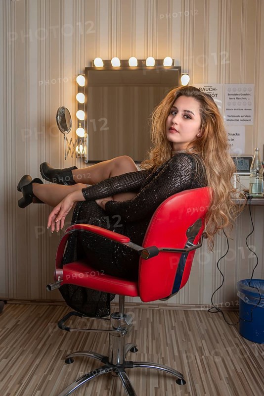 Young woman in black dress sits on a red chair in front of a dressing table