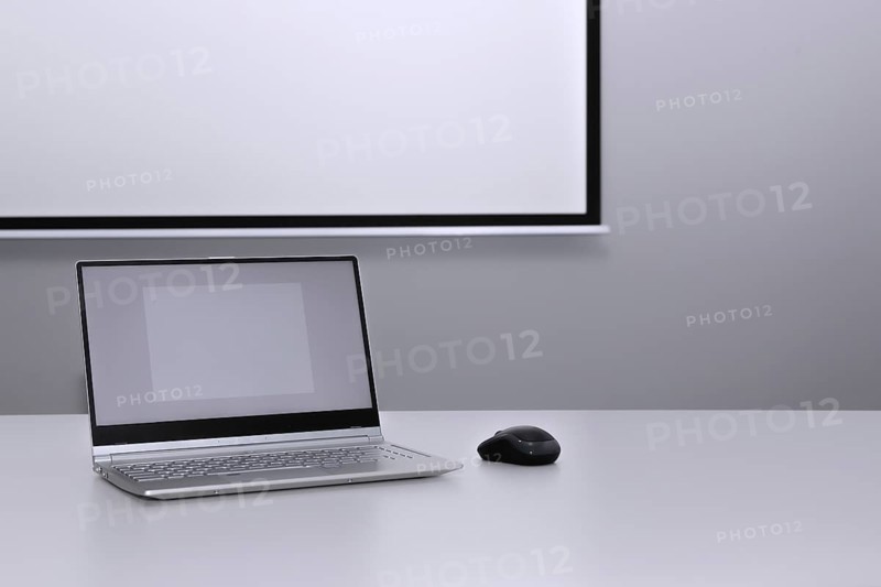 An unfolded laptop stands on a desk in front of a presentation screen
