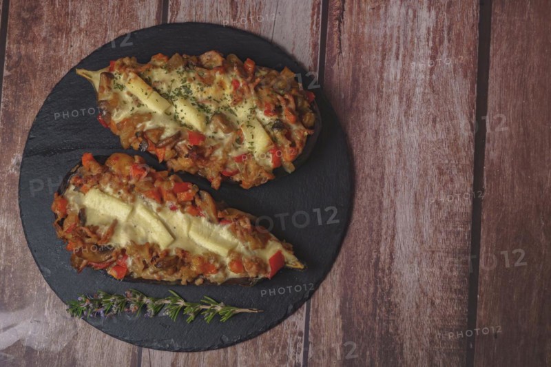 Foreground eggplant stuffed with meat and vegetables covered with bechamel sauce on a black slate on a wooden table