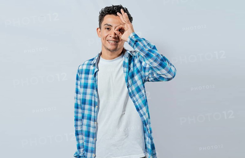 Happy young man making ok gesture with hand looking through fingers. Friendly man making ok gesture looking through fingers. Teenage guy making ok gesture looking through fingers