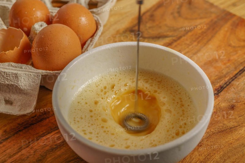 Beating an egg in a bowl with a small mixer with an egg cup at the bottom on a wooden table