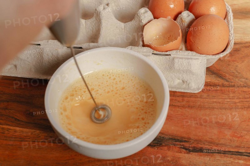Beating an egg in a bowl with a small mixer with an egg cup at the bottom on a wooden table