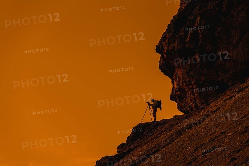 Landscape photographer in cliff face taking nature pictures of mountains with tripod at sunset in the Dolomites