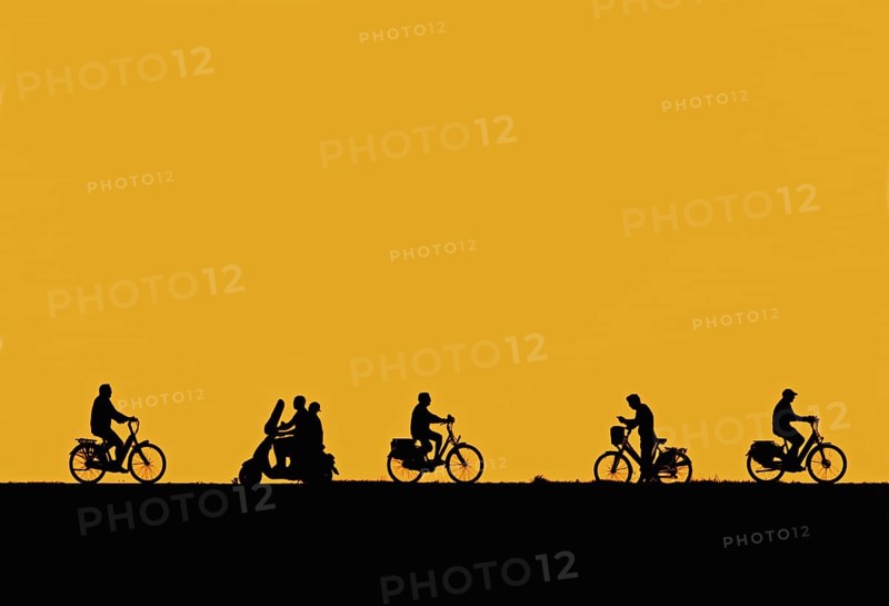 Motor scooter riding past cyclists and man on bicycle checking his smartphone silhouetted against orange sunset sky in summer