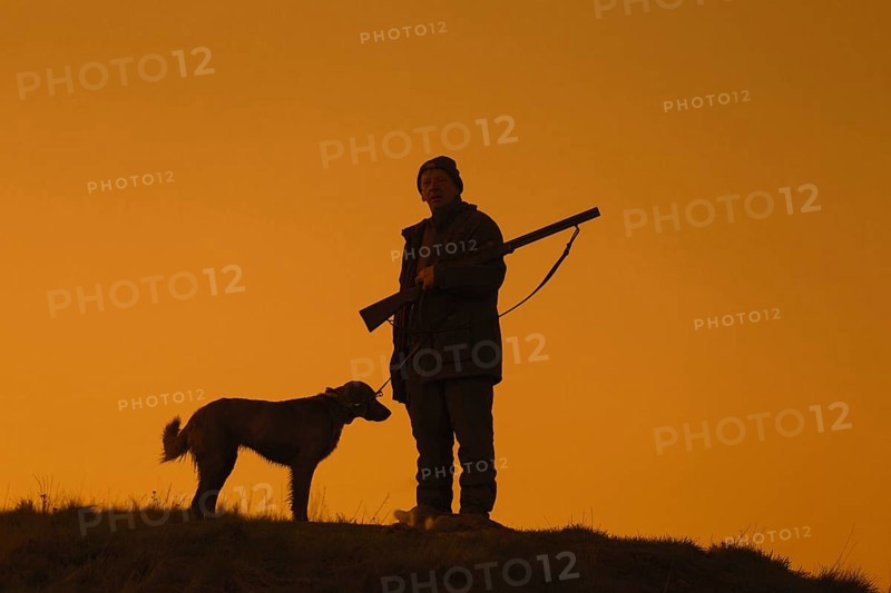 Hunter with hunting rifle and Weimaraner dog silhouetted against evening sky