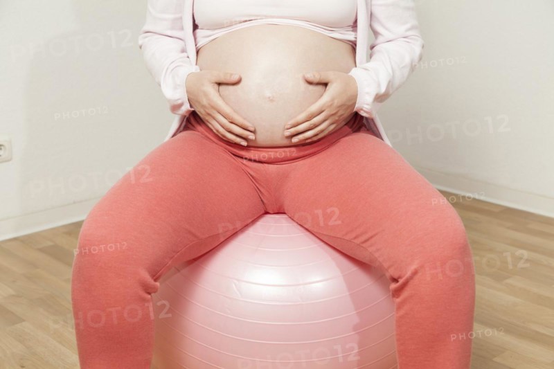 Pregnant woman sitting on exercise ball with hands on baby belly | MR:yes NH_pregnant_belly_mr