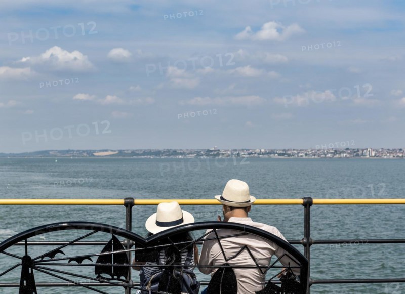Young Asian couple sitting on a bench ovelooking the sea wearing straw hats