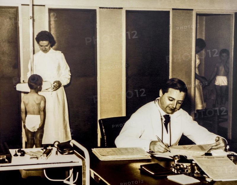Old sepia archival photograph showing doctor and nurse examining school children in their underwear at dispensary in the 1950s