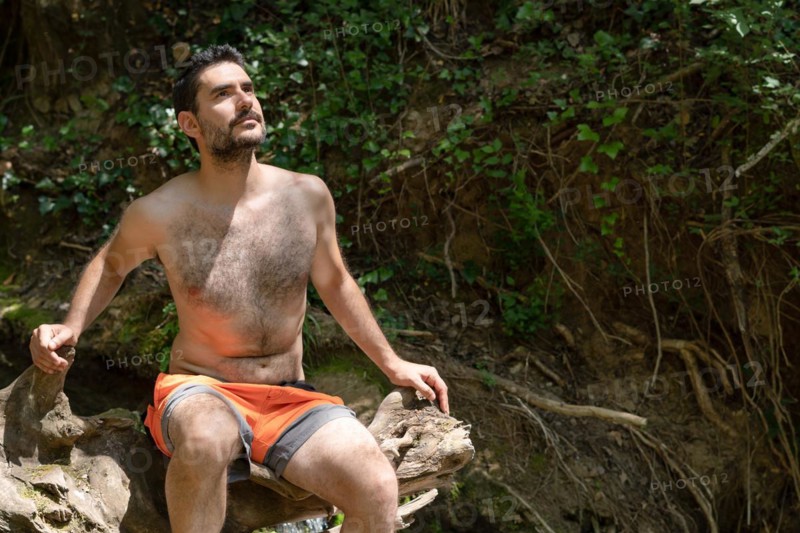 Man in swimsuit sitting on a tree trunk on a river bank